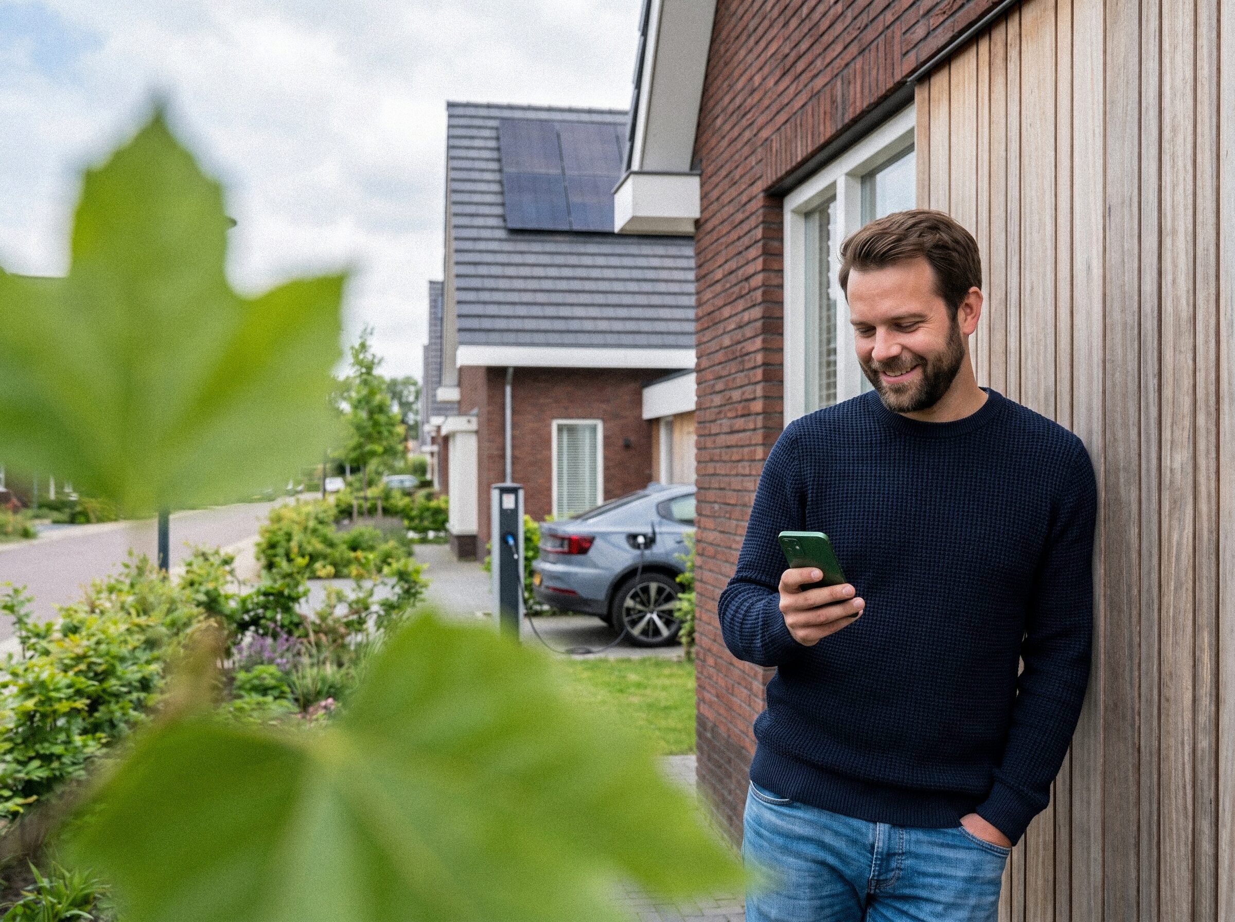 Man bekijkt zijn energieverbruik op zijn telefoon naast de laadpaal en thuisbatterij bij zijn woning